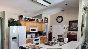 A kitchen with wooden cabinets, a refrigerator, a microwave, an oven, and a sink. There is a clock on the wall and a chandelier hanging above the sink.