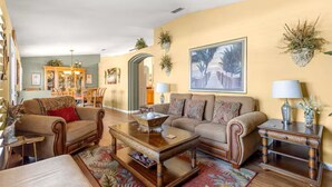 A living room with a sofa, armchairs, a coffee table, and a side table. There is a painting on the wall and a plant on the side table.