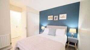 Een slaapkamer met een bed, twee nachtkastjes met lampen, een deur en drie ingelijste foto’s aan de muur.