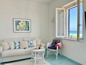 A room with a white sofa, a wicker chair, a small round table, a framed floral artwork, and a window with a view of the sea. - Charming rooftop apartment just 20 m from the sea with a fantastic sea view (Villanova Ostuni)