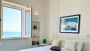 Una camera da letto con un letto grande, una finestra vista mare, un quadro appeso al muro e un comodino con un vaso verde.