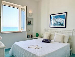 A bedroom with a large bed, a window with a view of the sea, a wall-mounted picture, and a bedside table with a green vase. - Charming rooftop apartment just 20 m from the sea with a fantastic sea view (Villanova Ostuni)