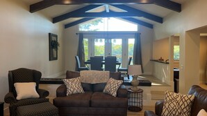 A spacious living room with a brown leather sofa, a patterned armchair, and a coffee table. A dining area with a table and chairs is visible through a glass door. - Deep water, private dock, beautiful sunsets, fishing, kayaking or boating. (Gainesville)