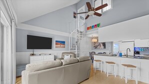 Ein modernes Wohnzimmer mit einer Couch, einem Fernseher, einem Deckenventilator und einer Küchenzeile mit Barhocker.