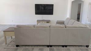 Ein Wohnzimmer mit einem grauen Ecksofa, einem kleinen Beistelltisch und einem an der Wand montierten Flachbildfernseher.