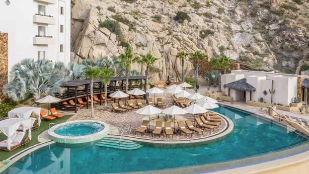 Ein Poolbereich mit Liegestühlen und Sonnenschirmen, umgeben von einer felsigen Landschaft und einem Gebäude mit Balkonen.