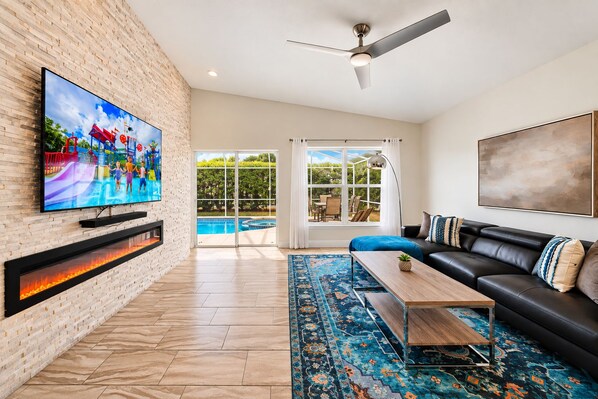 A modern living room with a flat-screen TV, a fireplace, a black leather sofa, and a large window overlooking a pool. - NEW LISTING Amazing Location! 4BR, Pool, Mid-Term Stays! Close to Disney! (Davenport)