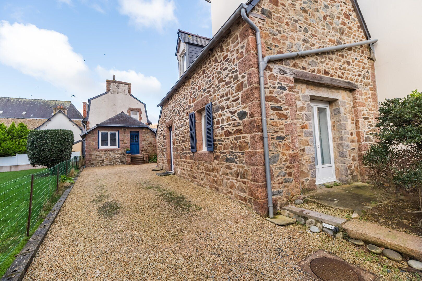 A stone building with a gravel driveway, a wooden fence, and a small garden area.