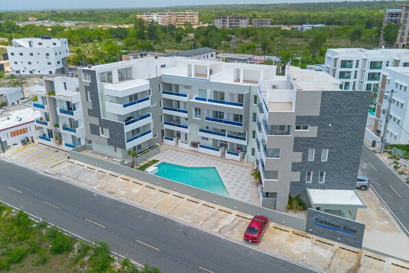 A modern multi-story hotel with a swimming pool, multiple balconies, and a parking area. - Caribbean Getaway 2BR at Caribbean Rose Bayahibe (San Rafael del Yuma)