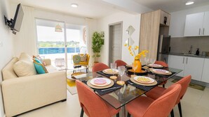 Una sala de estar moderna con un sofá, una mesa de comedor de vidrio para cuatro personas y una zona de cocina con armarios y un refrigerador.