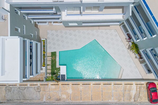Vue aérienne d’un bâtiment moderne doté d’une piscine, avec une voiture garée devant et une voiture rouge sur la droite.