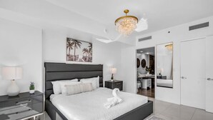 Ein modernes Schlafzimmer mit einem großen Bett, Nachttischen, einer Deckenleuchte und einem durch eine offene Tür einsehbaren Badezimmer.