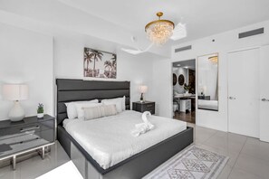A modern bedroom with a large bed, bedside tables, a chandelier, and a bathroom visible through an open door. - Miami Getaway | Icon Brickell 1BR in Downtown (Miami)