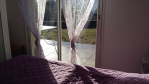 Een slaapkamer met een bed, een raam met dunne gordijnen en uitzicht op een patio.