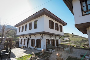 A two-story building with a white facade and brown wooden shutters. There are multiple benches and a small garden area with a stone pathway. - İkiz Konaklar (Şirince)