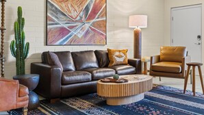 Een moderne woonkamer met een leren bank, een ronde houten salontafel, een cactus en een groot abstract schilderij aan de muur.