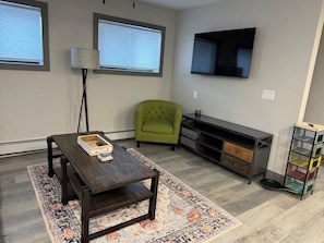A living room with a dark wooden coffee table, a green armchair, a TV mounted on the wall, and a pet cage. - 6th Ave Condo. Located in the center of town, right off Main Street. (Ouray)