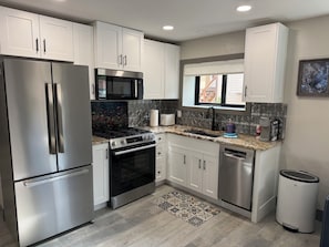 Cozinha moderna com eletrodomésticos de aço inoxidável, armários brancos, painel de fundo padronizado e janela com persianas.