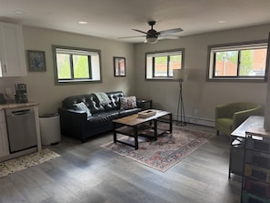 A living room with a sofa, coffee table, and a floor lamp. There are windows with blinds, a ceiling fan, and a kitchen area with a dishwasher and coffee maker. - 6th Ave Condo. Located in the center of town, right off Main Street. (Ouray)