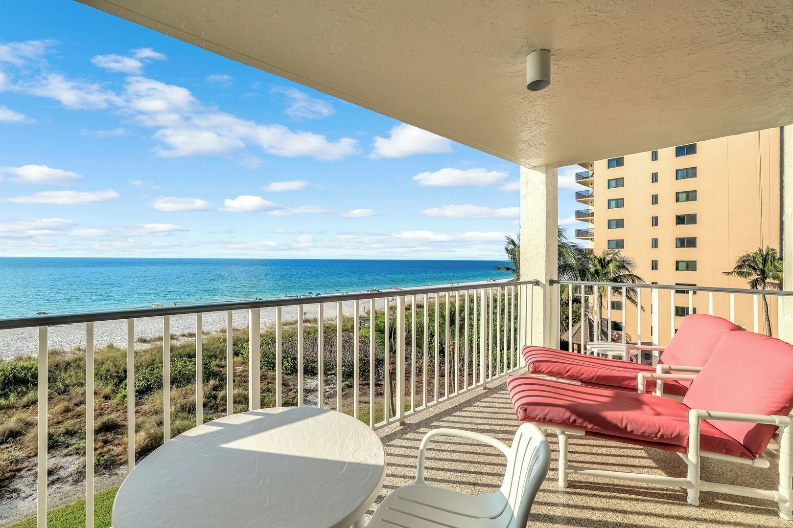 Un balcone con un tavolo bianco e due poltrone rosse imbottite, con vista su una spiaggia e l'oceano.