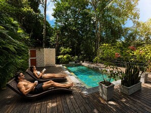 Un área junto a la piscina con dos sillones de descanso, un muro de piedra y abundante vegetación.
