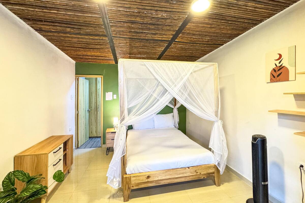 Una habitació amb un llit de fusta, una taula de nit, una planta i un ventilador de sostre.