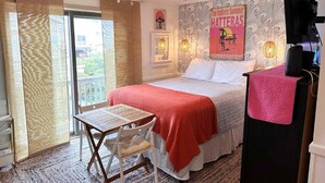 Une chambre avec un lit, une table et une chaise. On y trouve un ventilateur de plafond, un téléviseur et une affiche accrochée au mur.