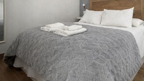Un letto rifatto con cura, coperta grigia a trama sfumata e asciugamani bianchi piegati, una testiera in legno e un comodino con una lampada.