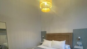 Un letto rifatto con una coperta grigia a trama decorativa e cuscini bianchi, un comodino con una lampada e un lampadario a sospensione sopra il letto.