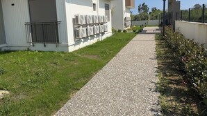 A residential building with a gravel pathway, a green lawn, and a row of air conditioning units on the exterior wall. - NestParkSide (Manavgat)