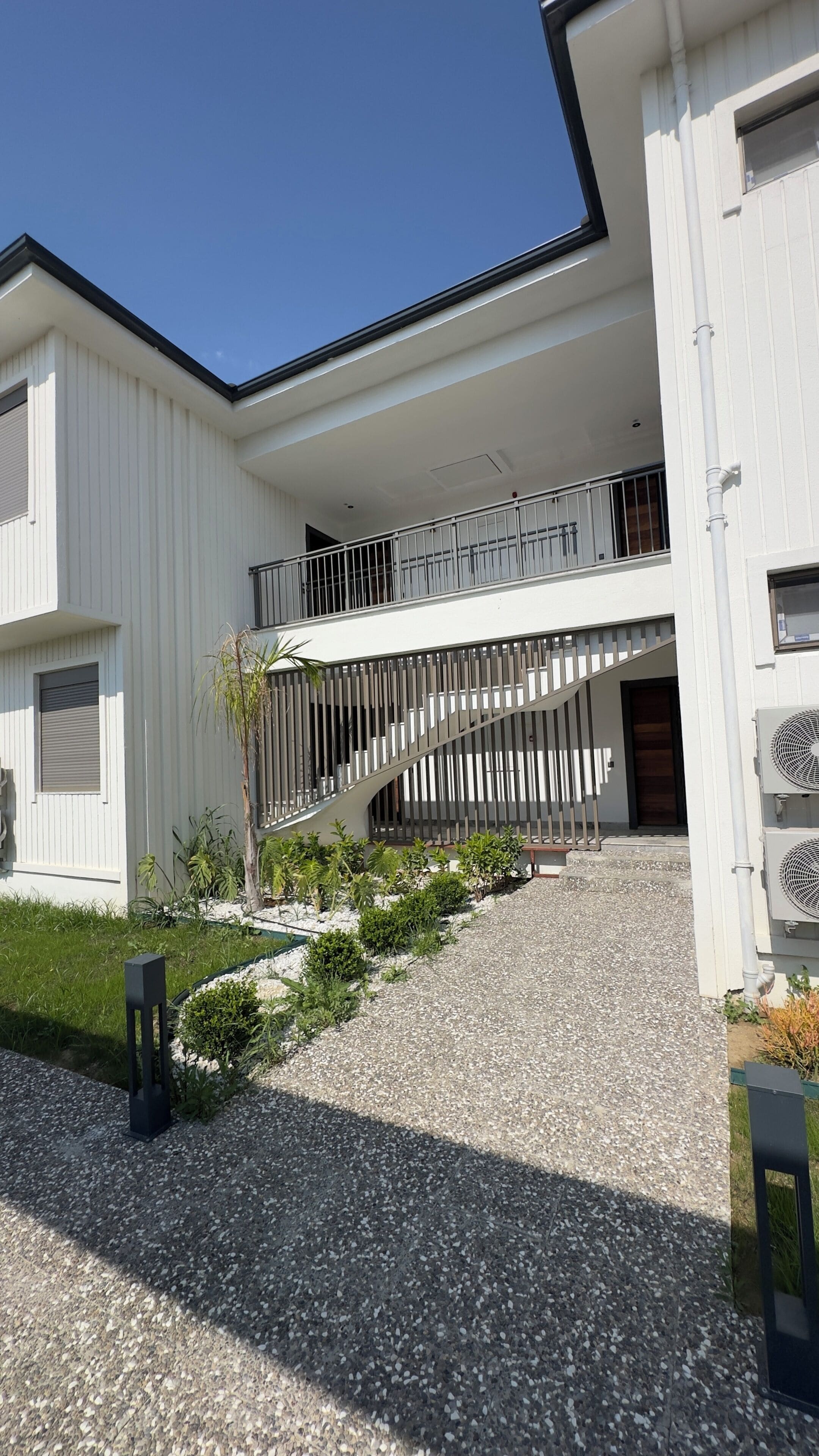Ein modernes Gebäude mit weißer Fassade, einer Schotterauffahrt und einer Treppe, die zu einem Balkon führt.