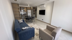 A modern living room with a sofa, a chair, a dining table with chairs, and a kitchen area with cabinets and a refrigerator. - NestParkSide (Manavgat)