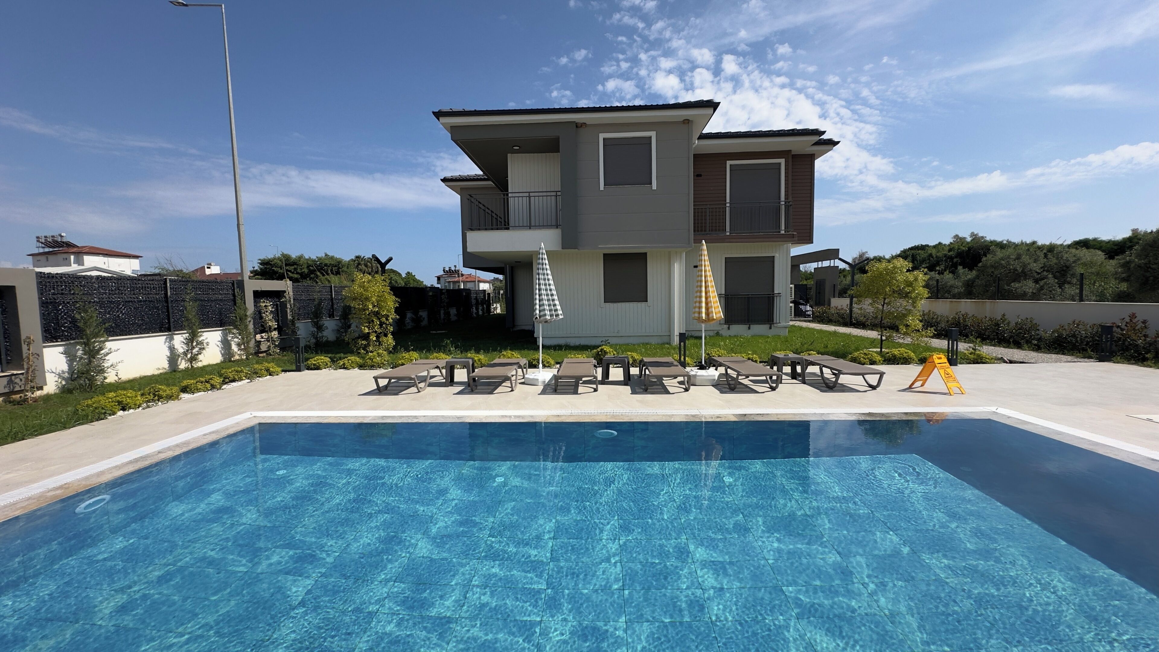 Ein Pool mit Liegestühlen und Sonnenschirmen, ein zweistöckiges Haus mit Balkon und ein klarer Himmel.