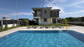 Une piscine avec des chaises longues et des parasols, une maison à deux étages dotée d’un balcon et un ciel dégagé.