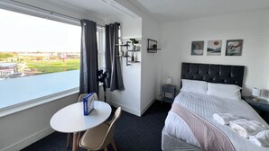 A hotel room with a bed, a small table with a chair, a bedside table with a lamp, a wall-mounted shelf with decorative items, and a window with curtains. - Triple room-Ensuite-Sea view (Skegness)