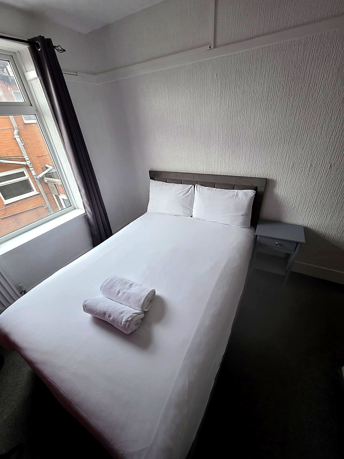 A neatly made bed with white linens and pillows, a small bedside table, and a window with curtains.