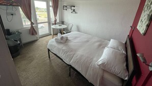 A hotel room with a bed, a table, and a chair. There is a window with curtains, a radiator, and a wall-mounted hanger. - Standard Triple Ensuite (Skegness)