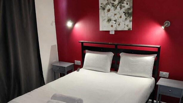 A hotel room with a bed, two bedside tables, a nightstand, a red wall, and a painting. - Standard Triple Ensuite (Skegness)