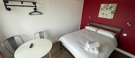 A hotel room with a bed, a table, a chair, a coat rack, and a painting on the wall.