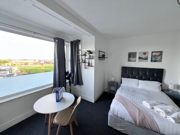 A hotel room with a bed, a small table with a chair, a bedside table with a lamp, a wall-mounted shelf with decorative items, and a window with curtains.
