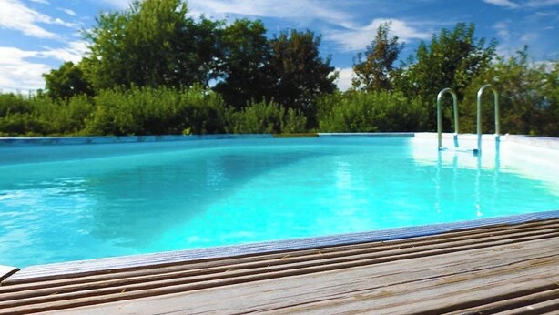 Una piscina con cubierta de madera y escalera de metal, rodeada de abundante vegetación.