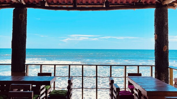 Um restaurante à beira-mar com mesas e bancos de madeira, telhado de chapa metálica e vista para o oceano.