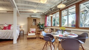 Um quarto espaçoso com piso de madeira, uma cama com roupa de cama vermelha e branca, uma mesa de jantar com cadeiras e grandes janelas com vista para o exterior.