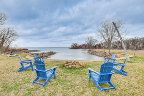 Un entorno junto al lago con cuatro sillas Adirondack azules dispuestas alrededor de un brasero, con vistas a un lago tranquilo.