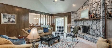 A cozy living room with a stone fireplace, wooden paneling, and a ceiling fan.