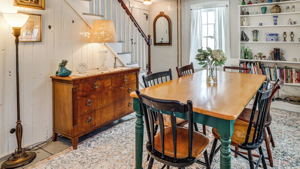 Un área de comedor con mesa y sillas de madera, una escalera, una estantería con adornos y una lámpara de pie.