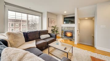 A living room with a fireplace, a sofa set, a coffee table, and a view of a cityscape through large windows.