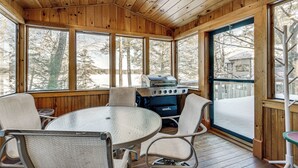 Una terraza de madera con una mesa redonda y sillas, una parrilla y grandes ventanales que ofrecen vistas a árboles cubiertos de nieve.