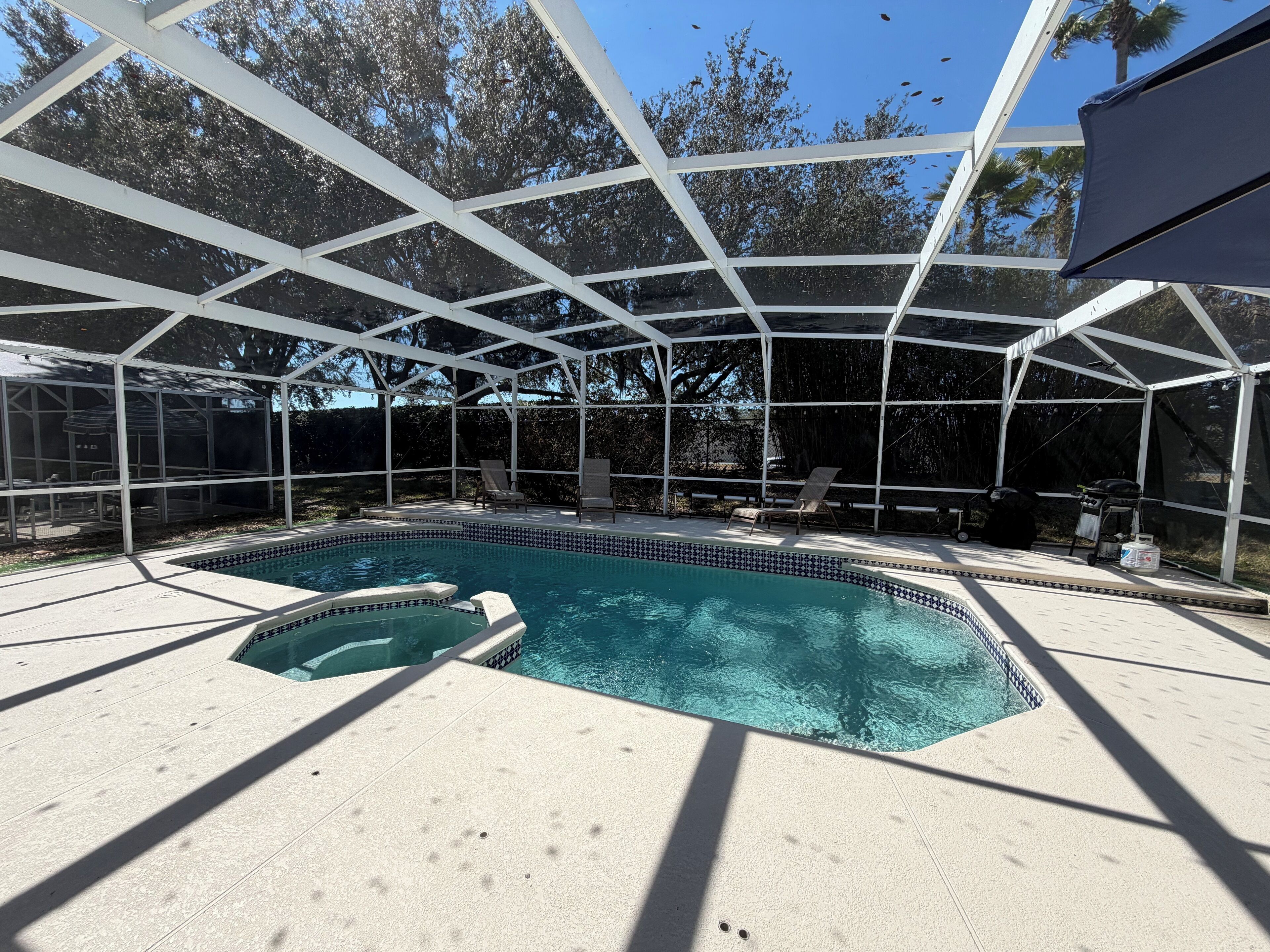 A covered outdoor swimming pool with a tiled edge, surrounded by a white pergola structure, and a patio area with lounge chairs.