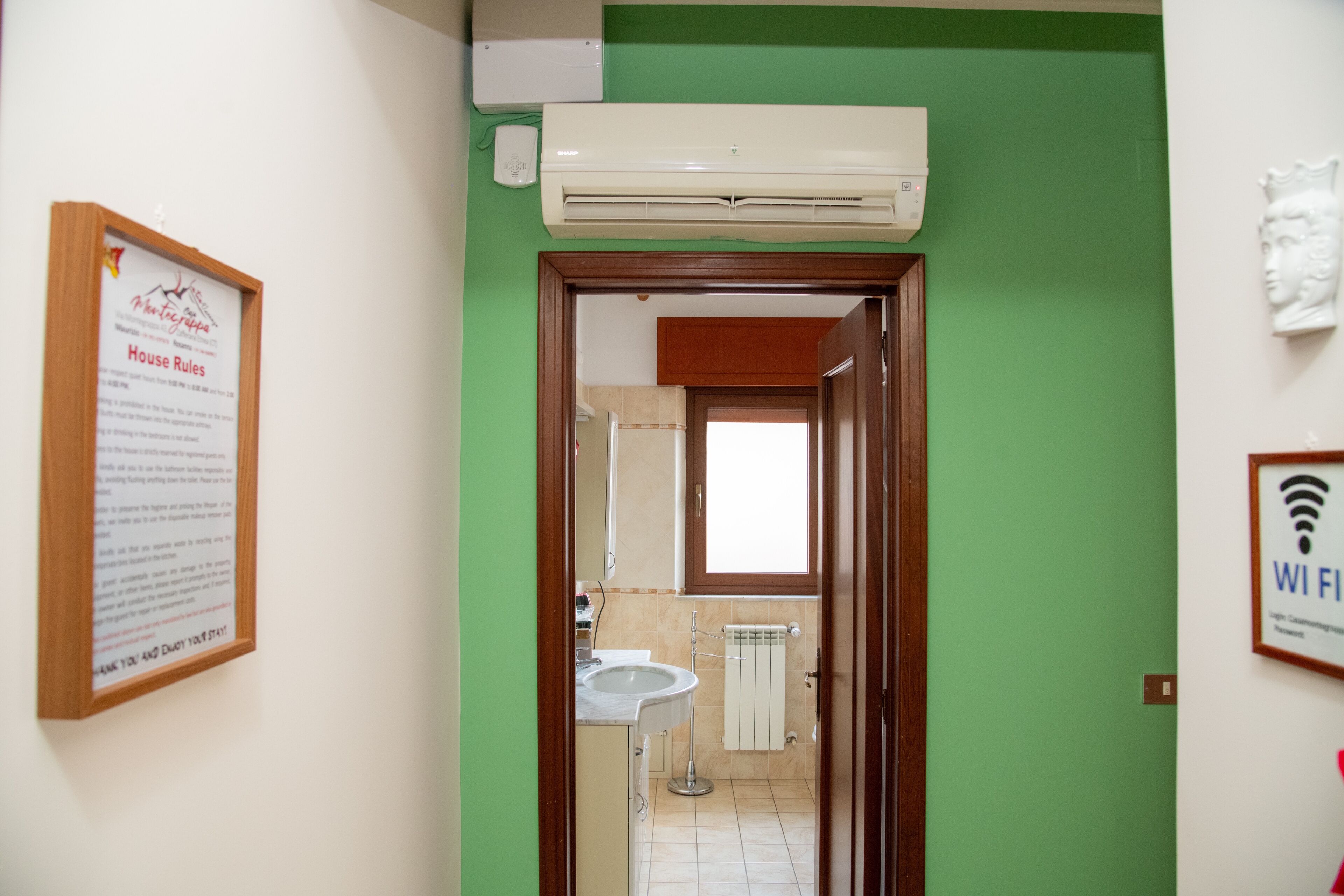 Une salle de bain avec un lavabo, un miroir et des toilettes. Un avis encadré est accroché au mur et un appareil de climatisation est installé au-dessus du lavabo.